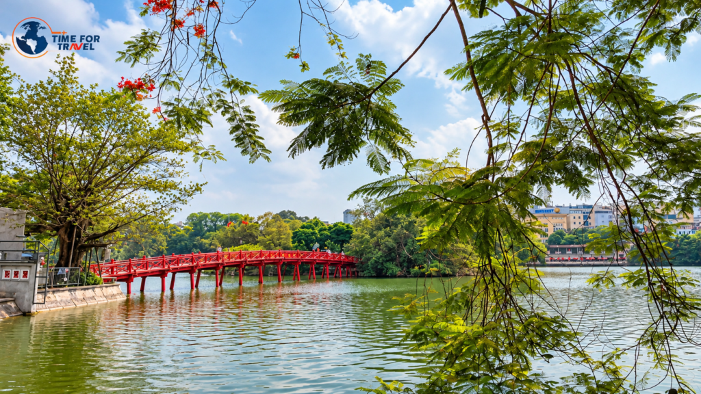 The iconic red Huc Bridge crossing Hoan Kiem Lake in Hanoi, Vietnam, surrounded by green trees.