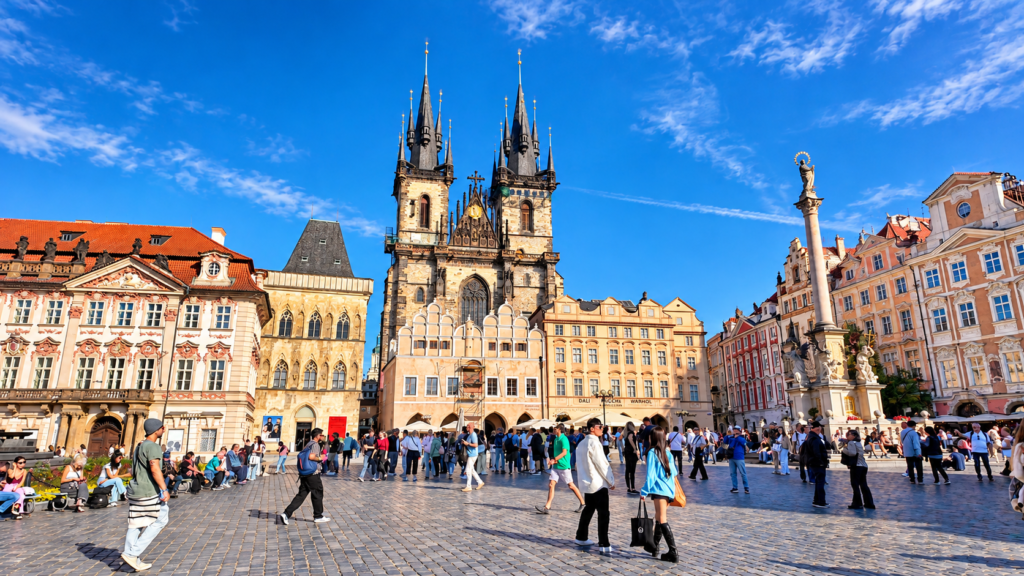 Tourists exploring Prague's Old Town Square, with the Gothic towers of the Church of Our Lady before Týn and colourful historic buildings.