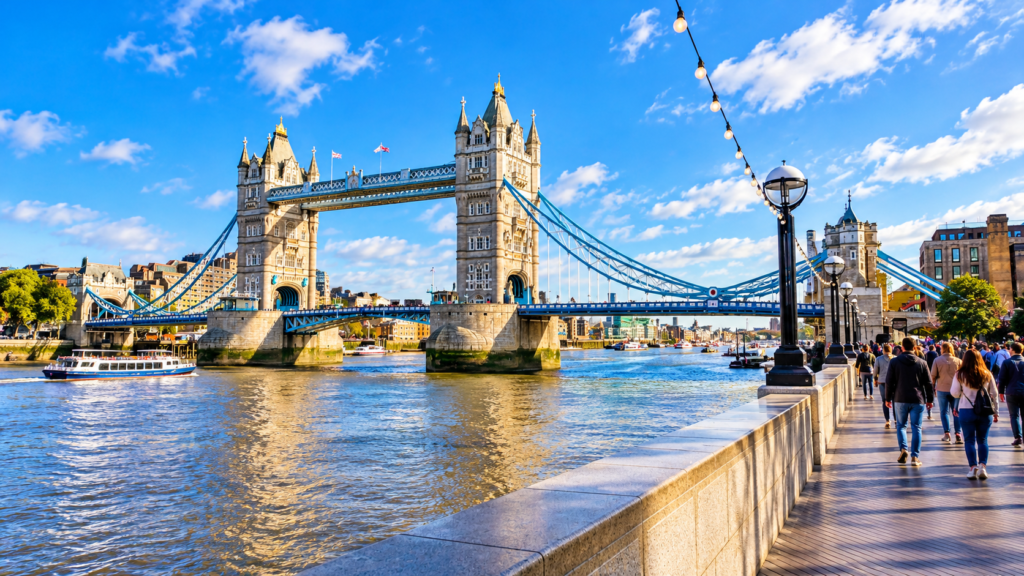 Tower Bridge spanning the River Thames in London, with its historic towers, blue suspension cables and people walking along the riverside.