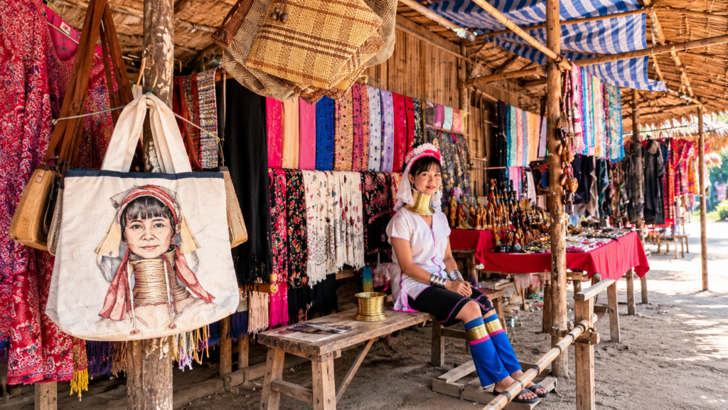 Kayan woman wearing traditional brass neck rings sitting beside colourful textiles and handmade souvenirs at the Long Neck Karen Village in Chiang Rai, Thailand.
