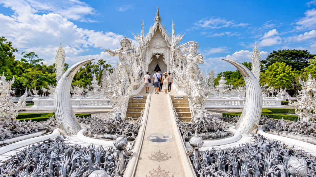 Visitors crossing the ornate white bridge at Wat Rong Khun (White Temple) in Chiang Rai, Thailand, surrounded by intricate sculptures and reaching hands.